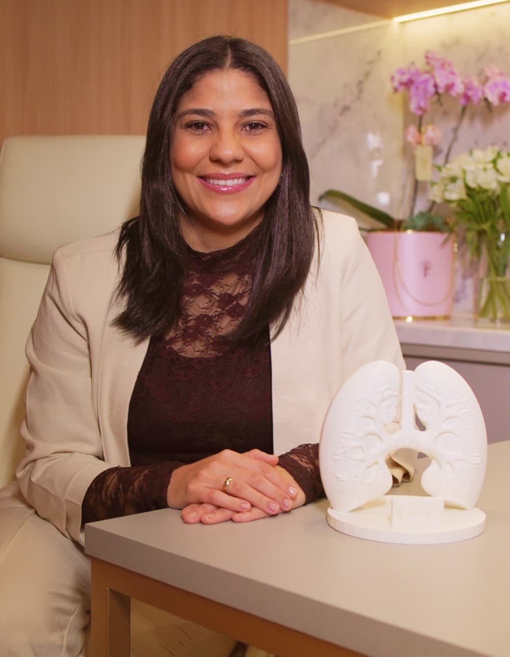Dra. Camilla Matos, cirurgiã torácica, sorrindo ao lado de um modelo anatômico de pulmões em seu consultório em Feira de Santana